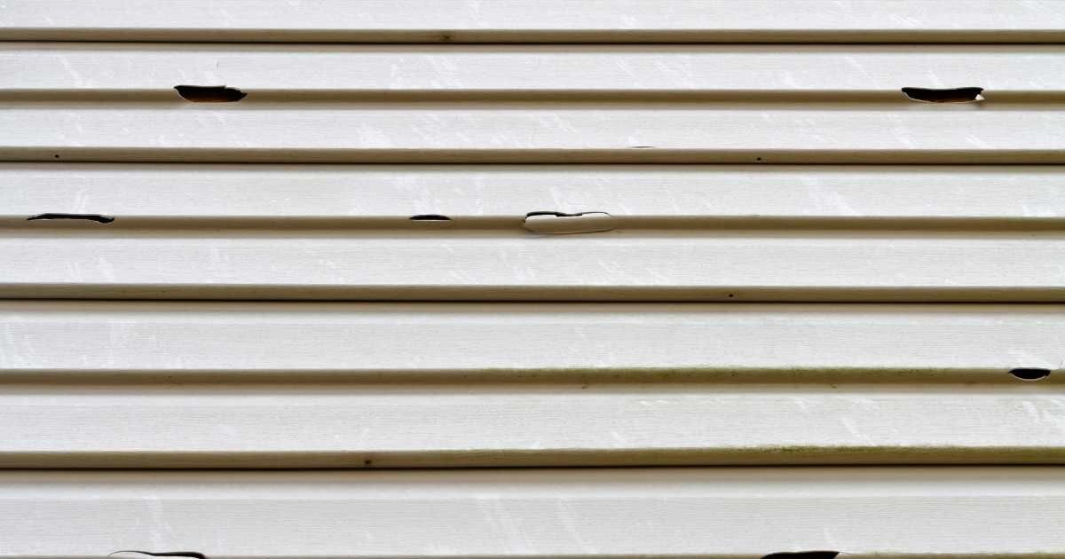 A close-up of beige vinyl siding with several visible cracks and damages.